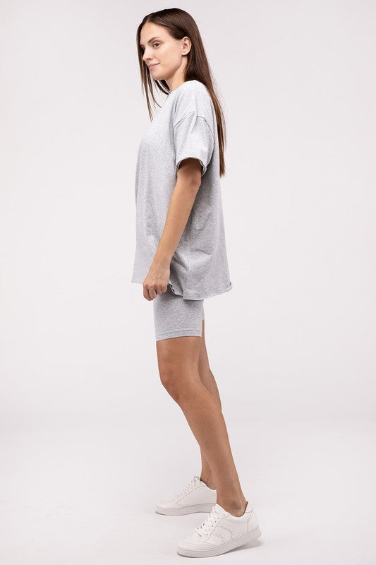 A woman stands in a studio, wearing a loose grey cotton round neck top & biker shorts set, and white sneakers, posing with one leg slightly bent.