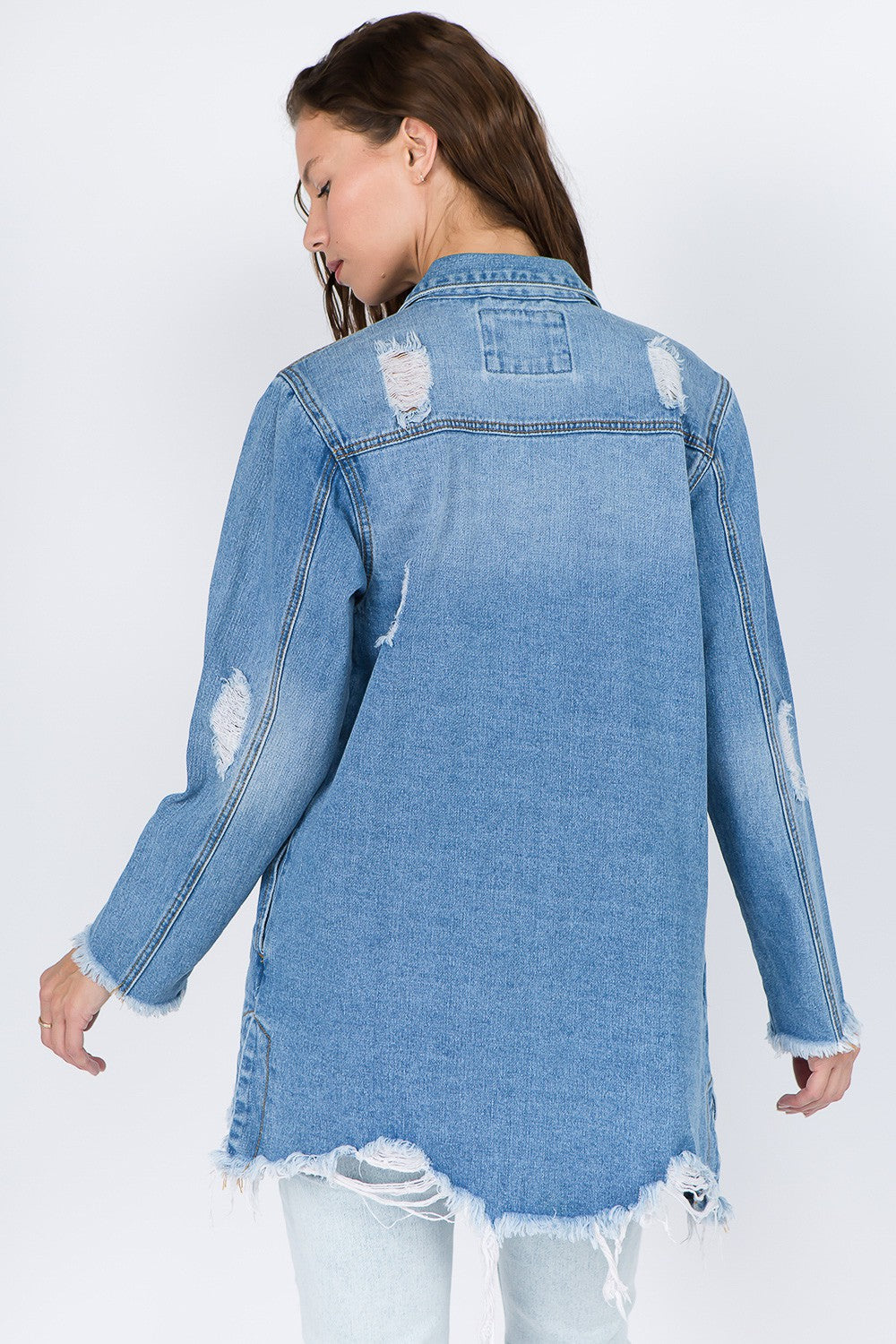 A woman models the American Bazi Distressed Frayed Hem Denim Jacket, featuring frayed edges, a button-up front, and chest pockets that add to its vintage feel. She has long hair and is looking downward with one hand touching her head.