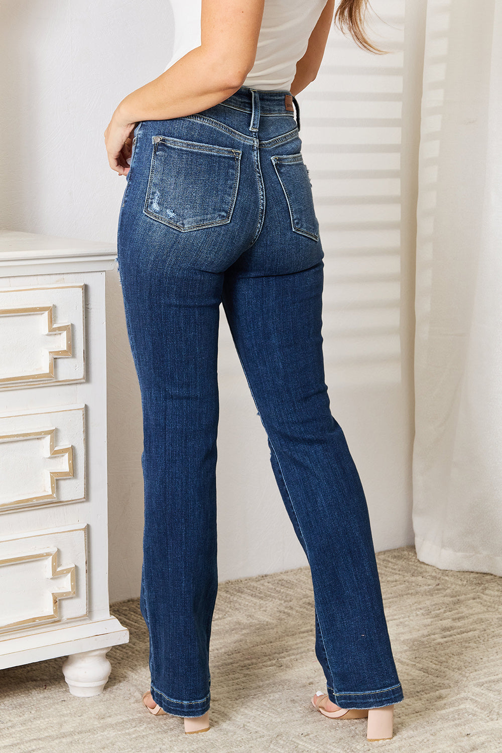 A person wearing Judy Blue Full Size Mid Rise Hand Sand & Destroy Bootcut Jeans stands with hands in pockets. The slightly stretchy jeans have rips on both legs, and the person is standing indoors beside a white dresser.