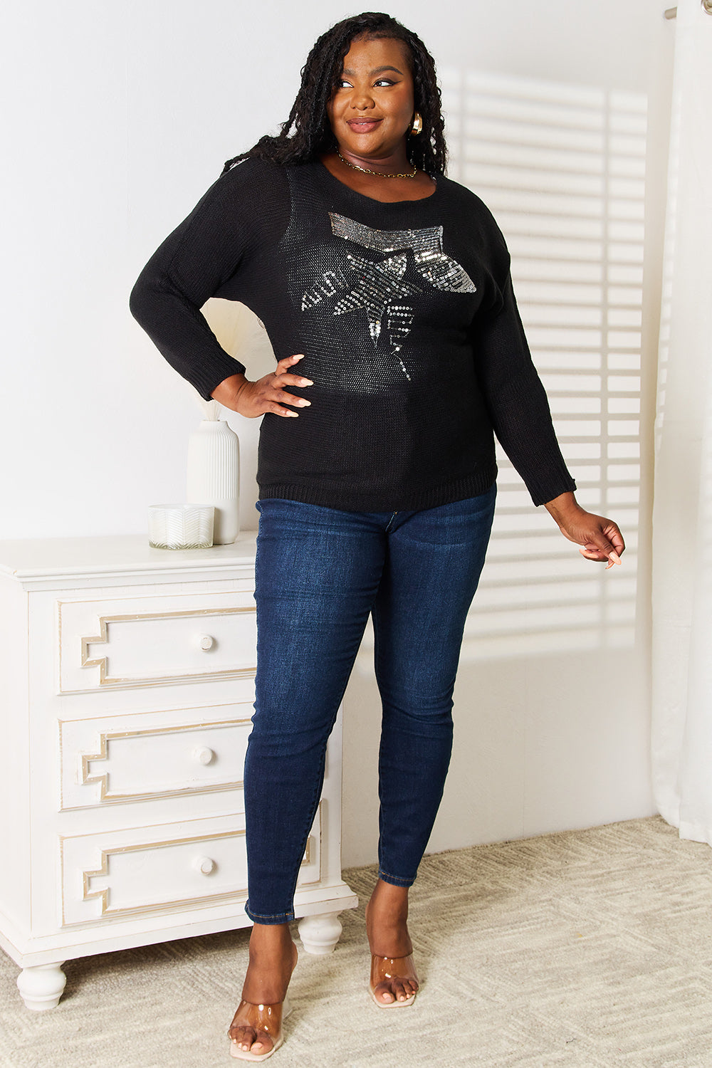 A person wearing a Sequin Graphic Dolman Sleeve Knit Top paired with blue jeans stands near a white dresser adorned with decor items.