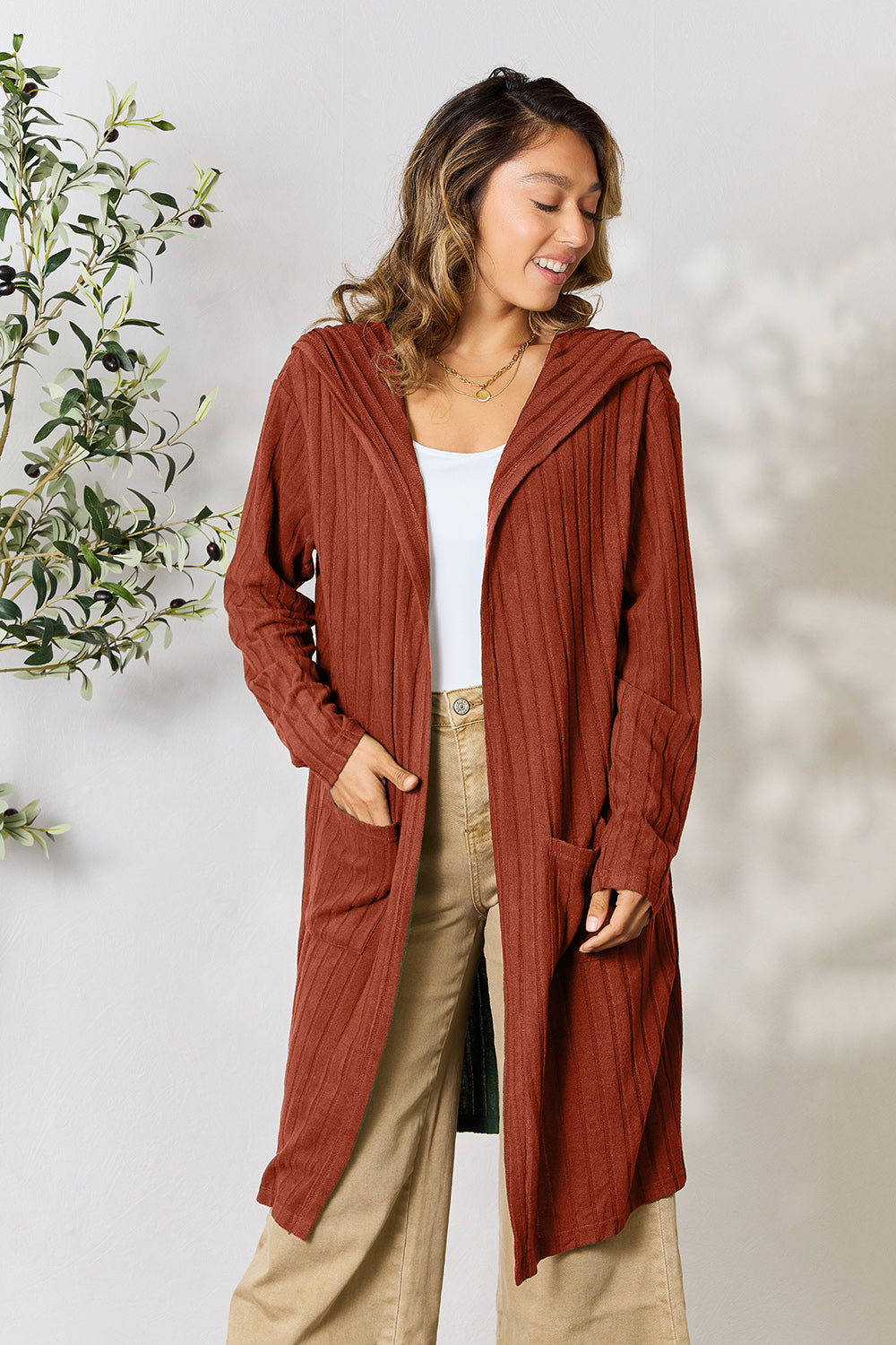 A woman wearing the Basic Bae Full Size Hooded Sweater Cardigan in black, paired with a white top and khaki pants, stands in front of a light background accented by a leafy plant on her left. With one hand tucked into her pocket, she gazes to her right.