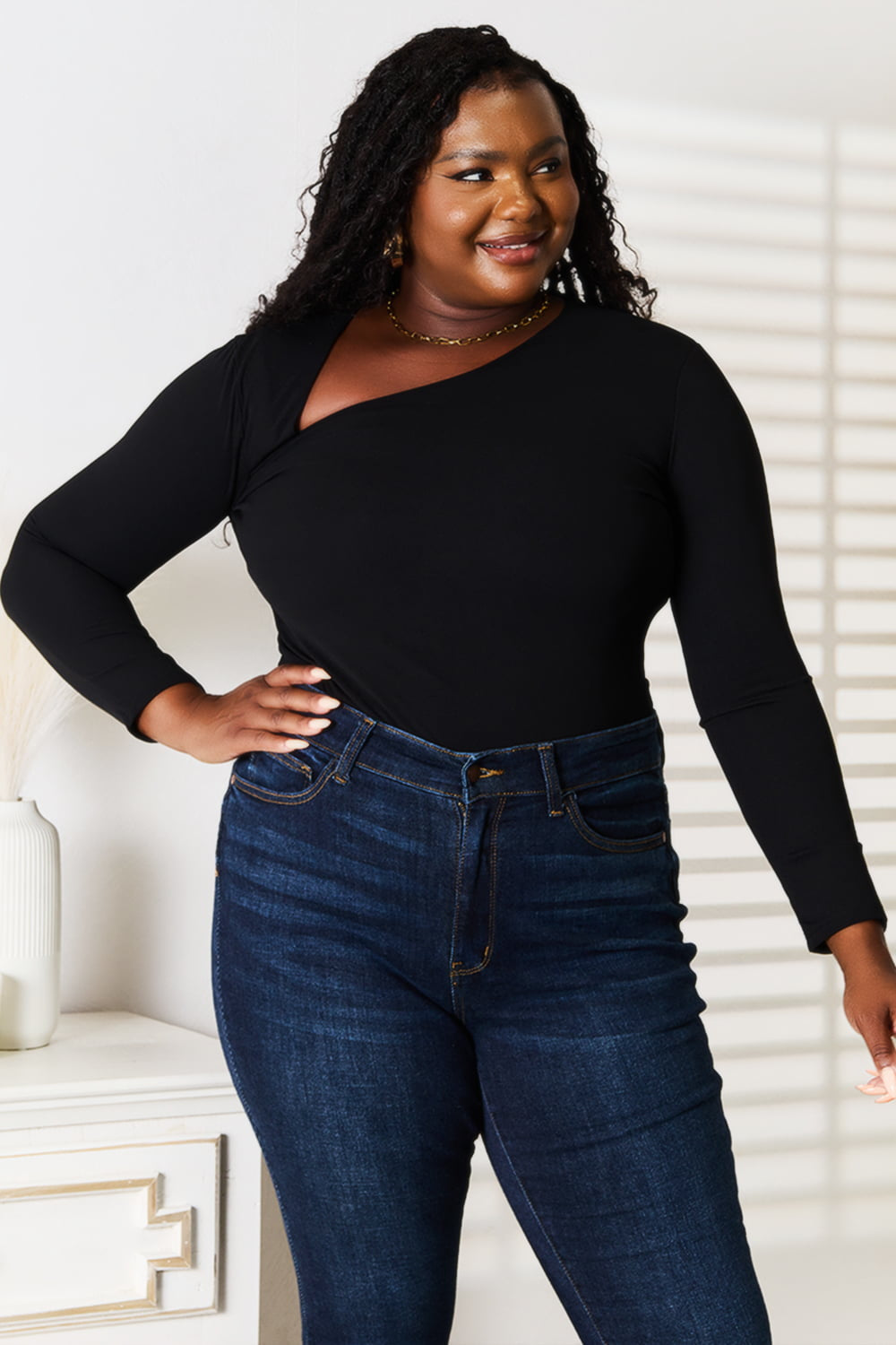 A woman with long hair is wearing a modern, stylish wardrobe featuring the Basic Bae Asymmetrical Neck Long Sleeve Top and jeans. She stands indoors in front of a white wall with shadow patterns.