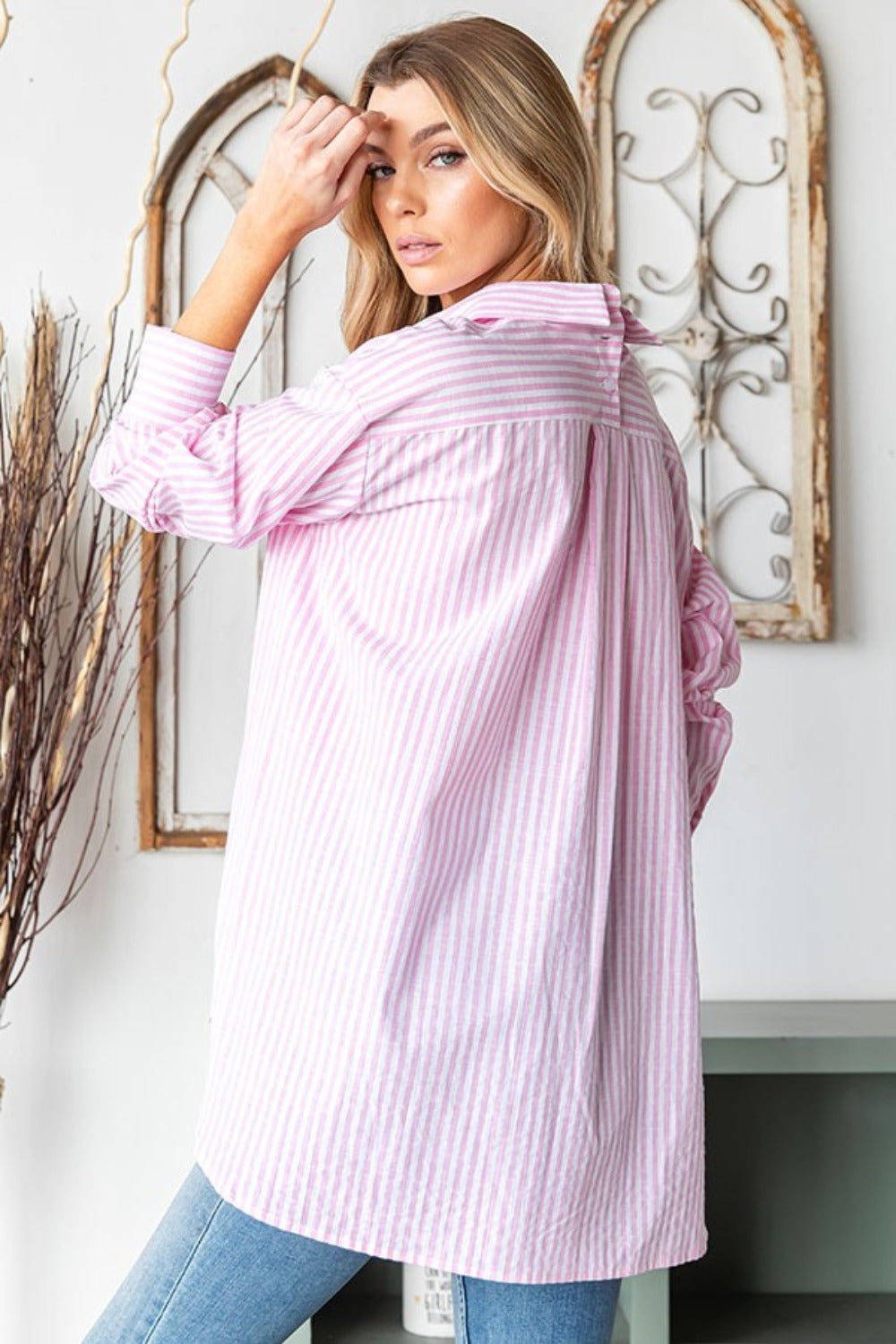 A woman wearing the First Love Full Size Striped Button Down High-Low Hem Shirt in pink and white, paired with blue jeans, stands in front of a decorative background, posing with one hand touching her hair.