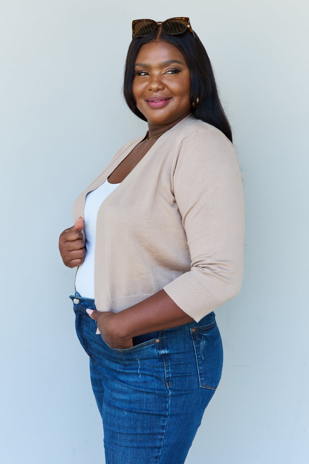 Woman with long dark hair wearing the versatile Doublju My Favorite Full Size 3/4 Sleeve Cropped Cardigan in Khaki over a white top and blue jeans, standing against a plain background.