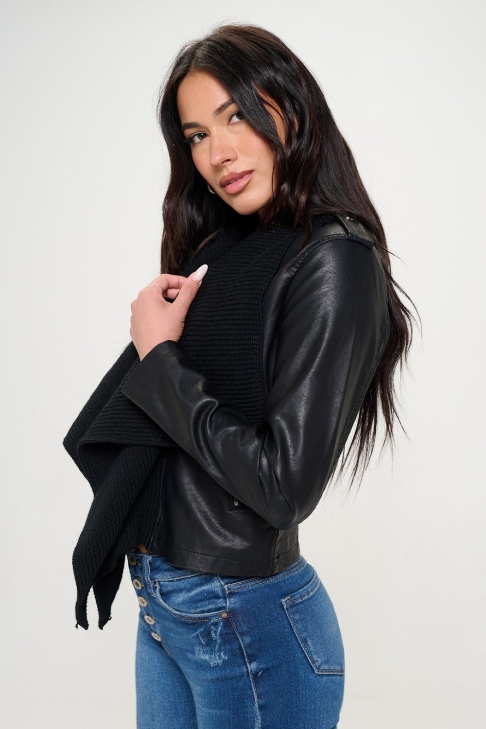 A woman with long, dark hair wearing a black top, a black shawl over the Coalition LA Knit Collared Faux Leather Crop Jacket, and blue jeans stands against a plain white background.