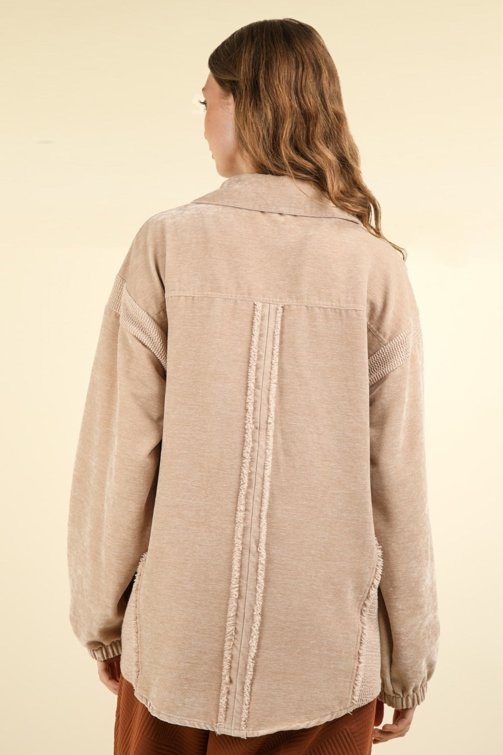 A woman with long wavy hair wears the VERY J Mixed Media Button Down Raw Hem Shacket featuring a drop shoulder design over a brown outfit. She faces the camera with one hand holding the lapel of her shacket against a plain beige background.