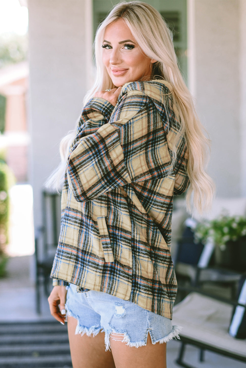 A person with long blonde hair, seen from the back, wearing a Khaki Plaid Button Neck Pocketed Pullover Hoodie and denim shorts, stands on a porch.