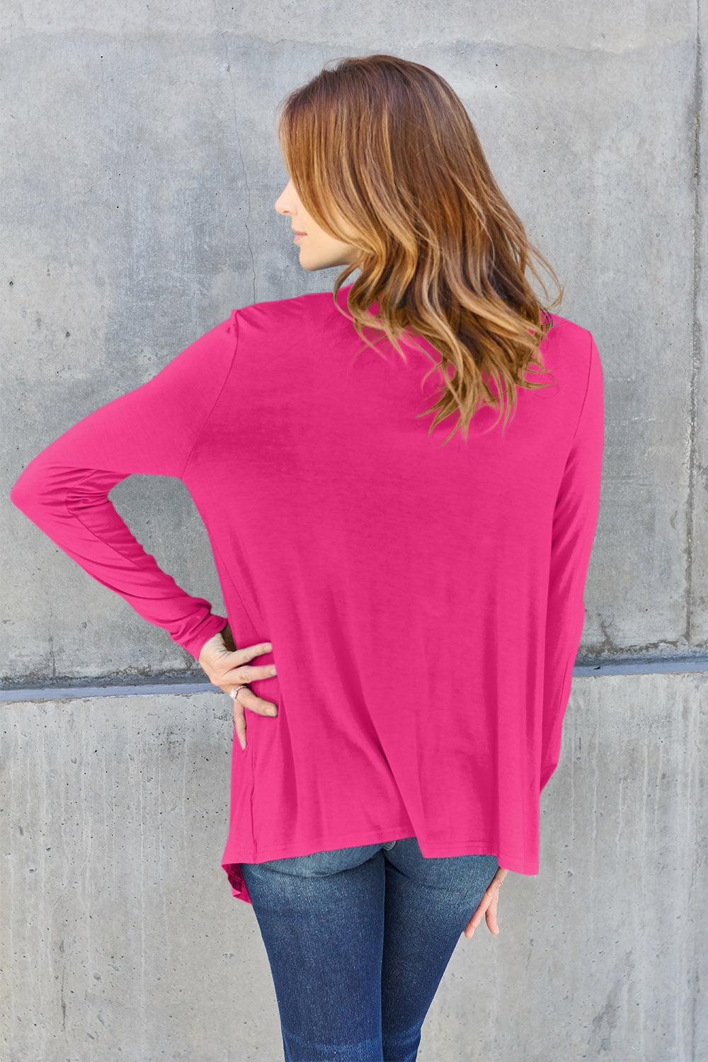 A woman wearing the Basic Bae Full Size Open Front Long Sleeve Cardigan in navy blue, paired with a white top and blue jeans, stands against a concrete wall. She touches her hair and gazes to the side, embodying a chic yet simple style. The slightly stretchy materials of the cardigan ensure comfort while maintaining its stylish simplicity. For easy care, machine wash it cold.