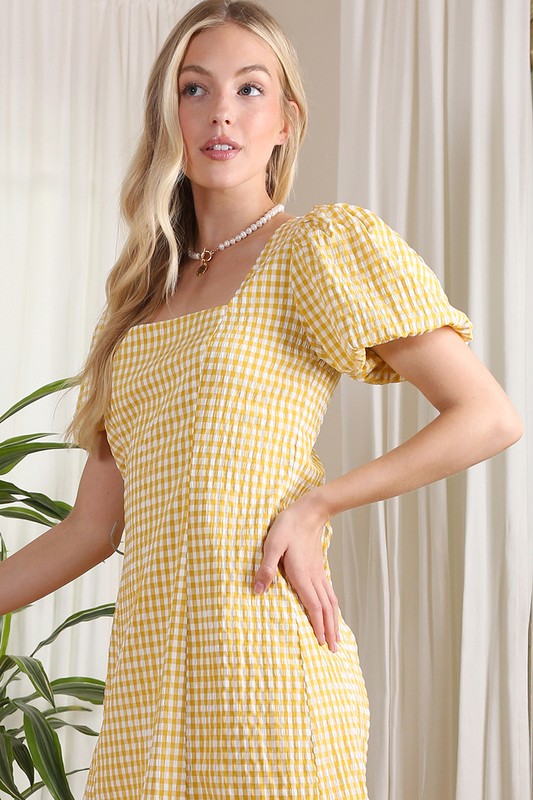 A woman stands indoors, smiling while wearing the SS back strap dress in yellow gingham, and holding a white purse.