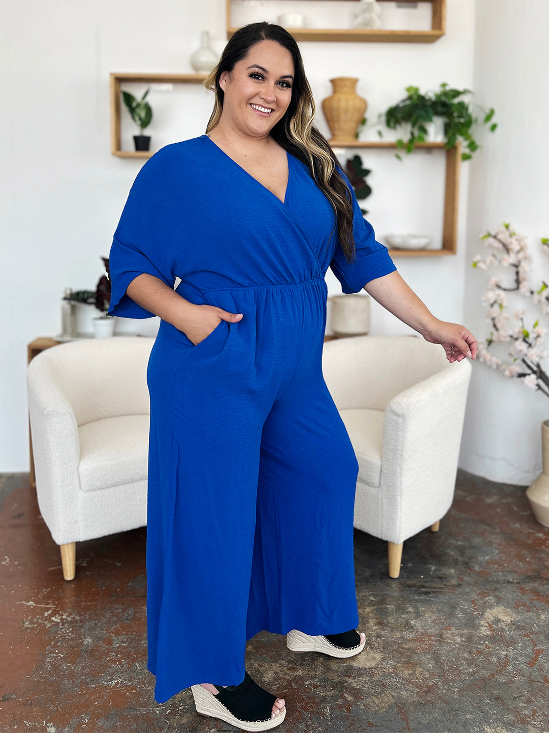 Person wearing the Double Take Full Size Surplice Wide Leg Jumpsuit with Pockets in royal blue, featuring wide legs, a deep V-neckline, and ruched detailing, posing with hands on hips against a plain light-colored background.