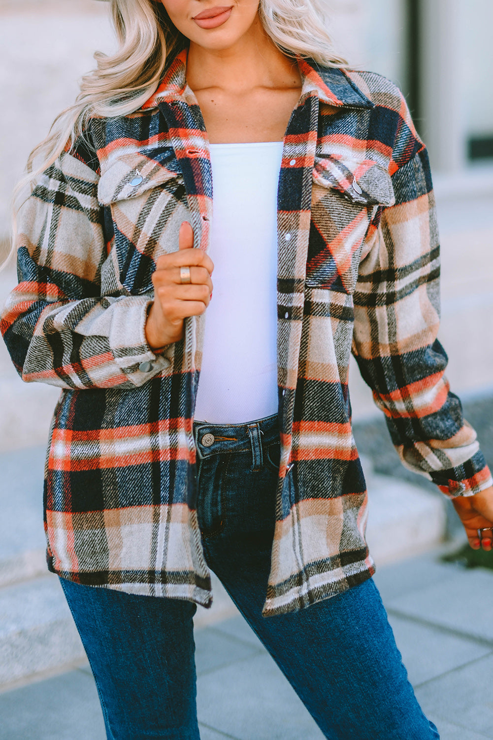 A person wearing a Brown Plaid Button Up Long Sleeve Flannel Shacket layered over a white top with ripped jeans and holding a brown handbag stands against a textured gray wall.
