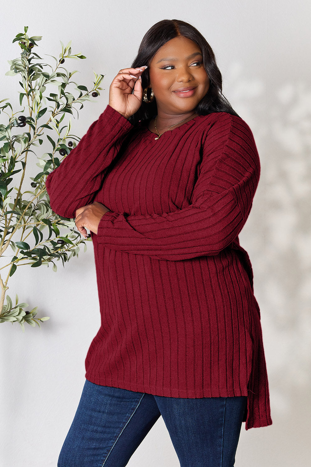 A person with shoulder-length hair smiles while wearing a beige ribbed sweater called the Basic Bae Full Size Ribbed Round Neck Long Sleeve Slit Top, along with matching beige pants, showcasing a basic style. They stand next to a green plant.