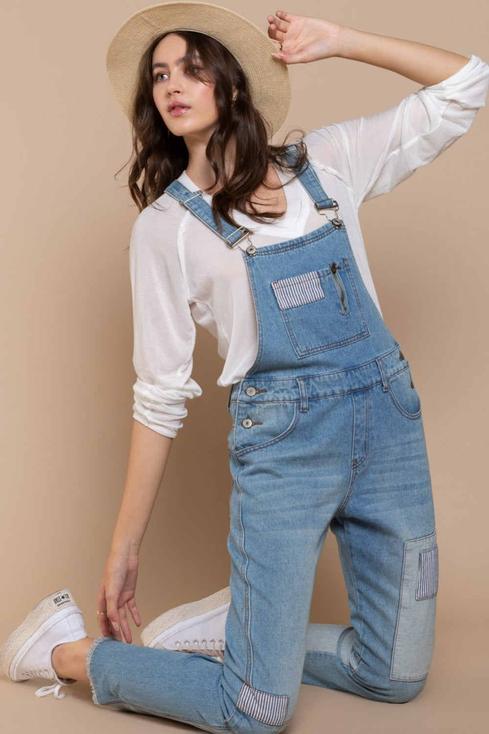 A woman kneels on one knee against a beige background, wearing a beige hat and white long-sleeve shirt, paired with POL Front Chest Zipper Slim Leg Denim Overalls featuring patches.