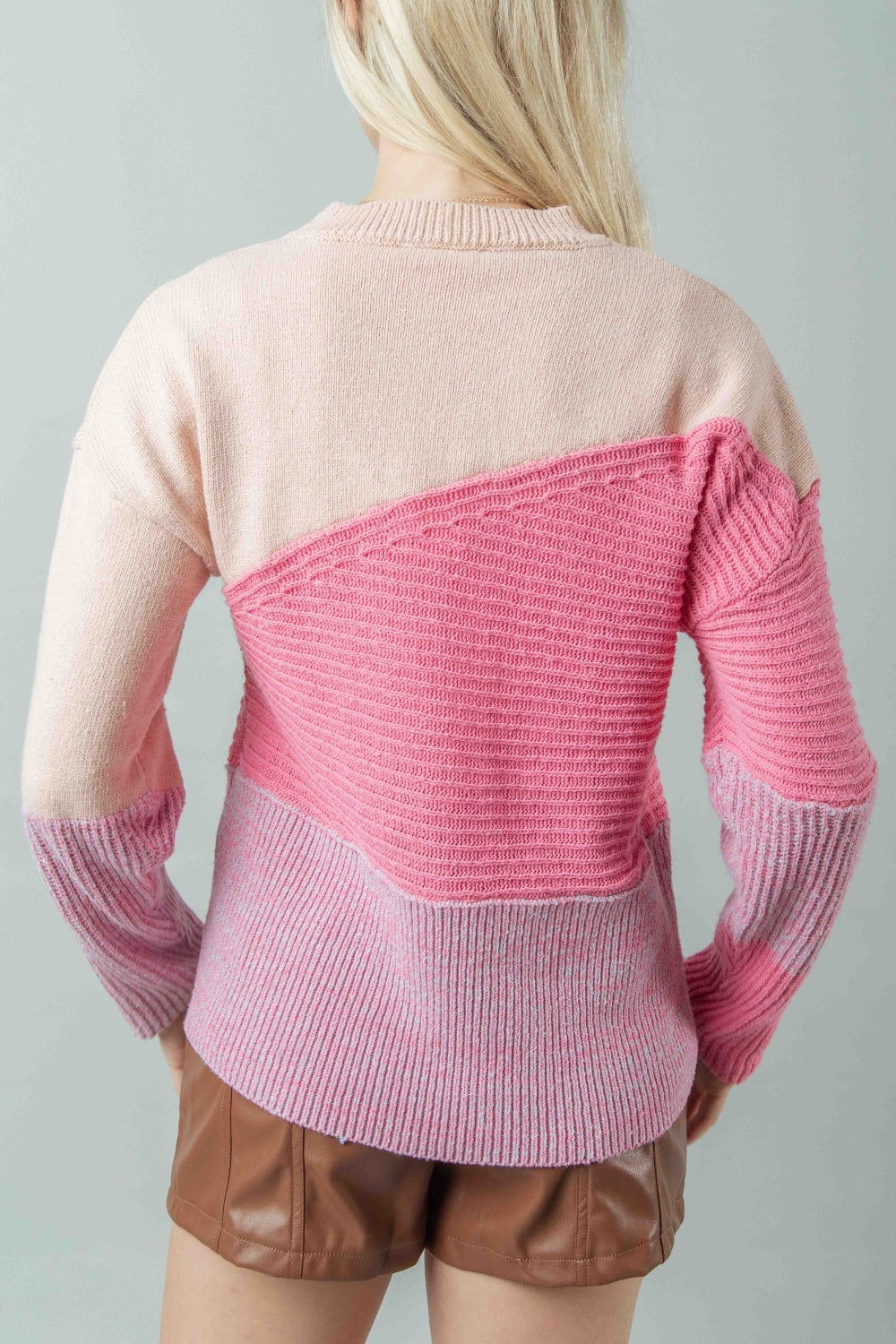 A woman stands against a plain background, elegantly dressed in a pink VERY J Color Block Long Sleeve Sweater and brown shorts, showcasing a chic blend of style and comfort.