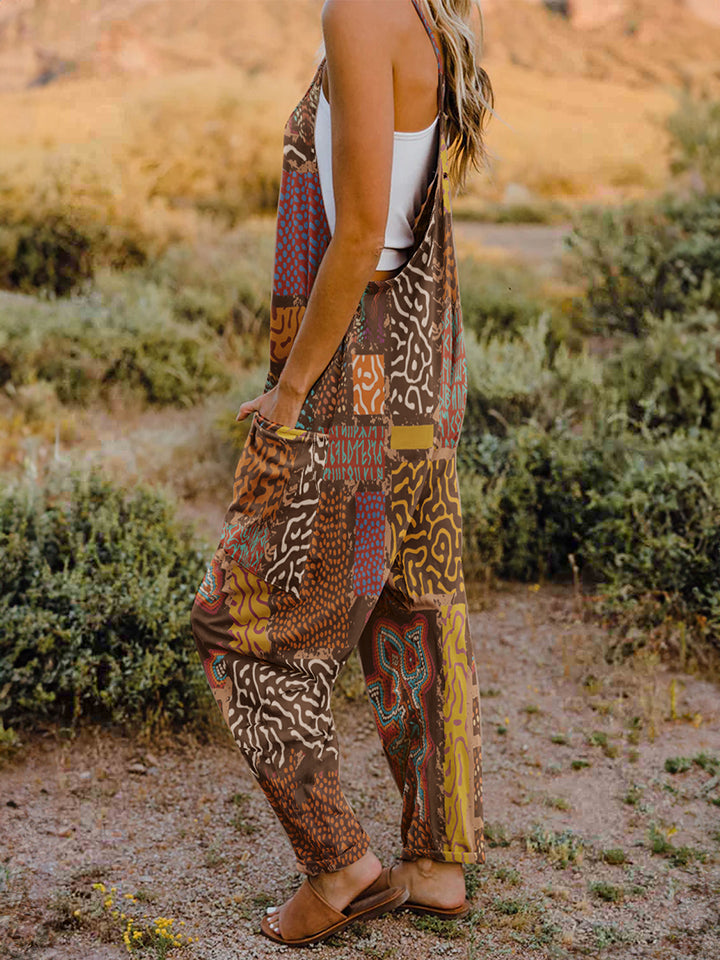 A person standing outdoors in front of rocky terrain, wearing a Full Size Printed V-Neck Sleeveless Jumpsuit and a hat.