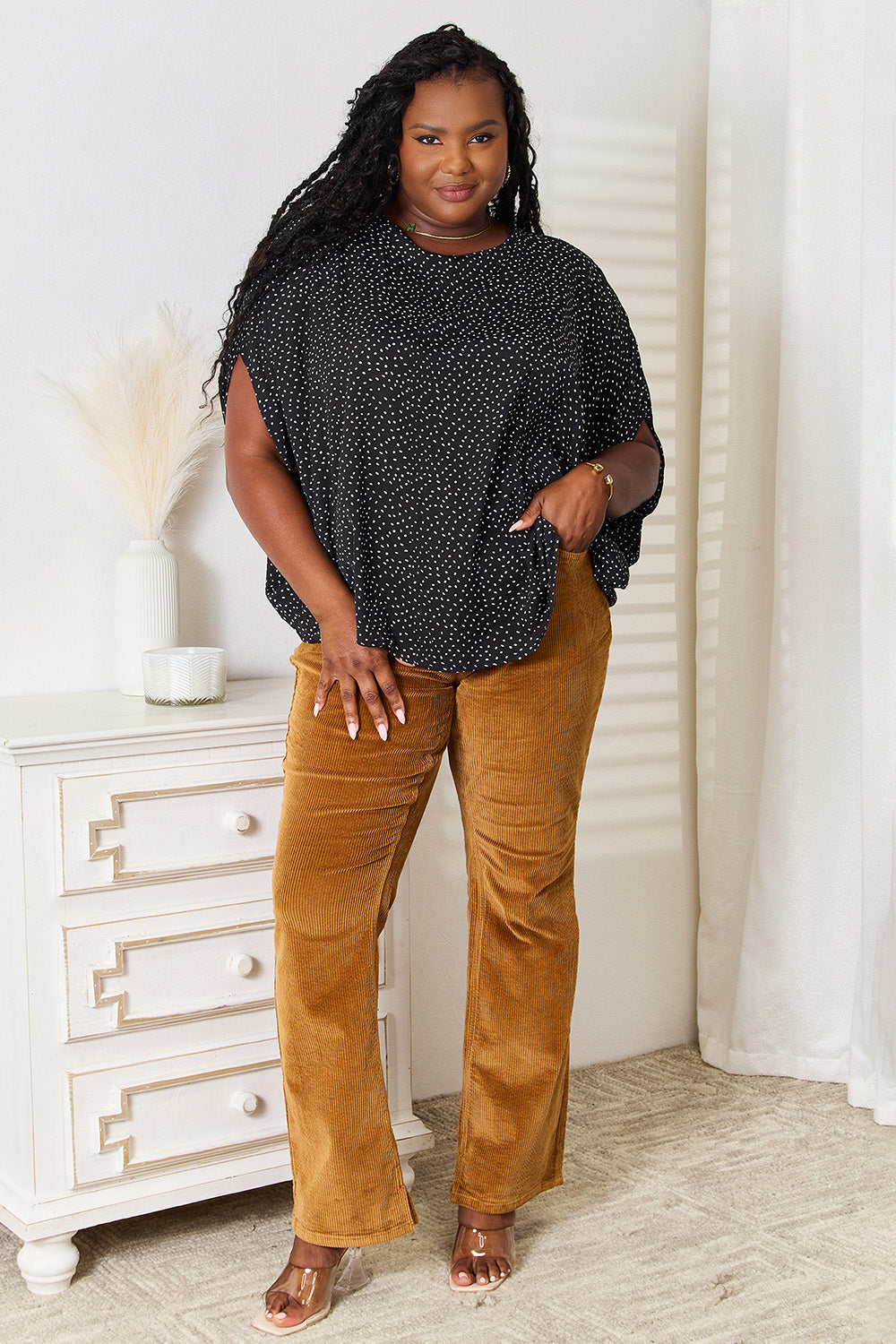 A woman stands wearing the Perfee Printed Dolman Sleeve Round Neck Blouse with white dots and black pants, smiling and looking to her left. The blouse features a printed design that adds a touch of elegance to her outfit.