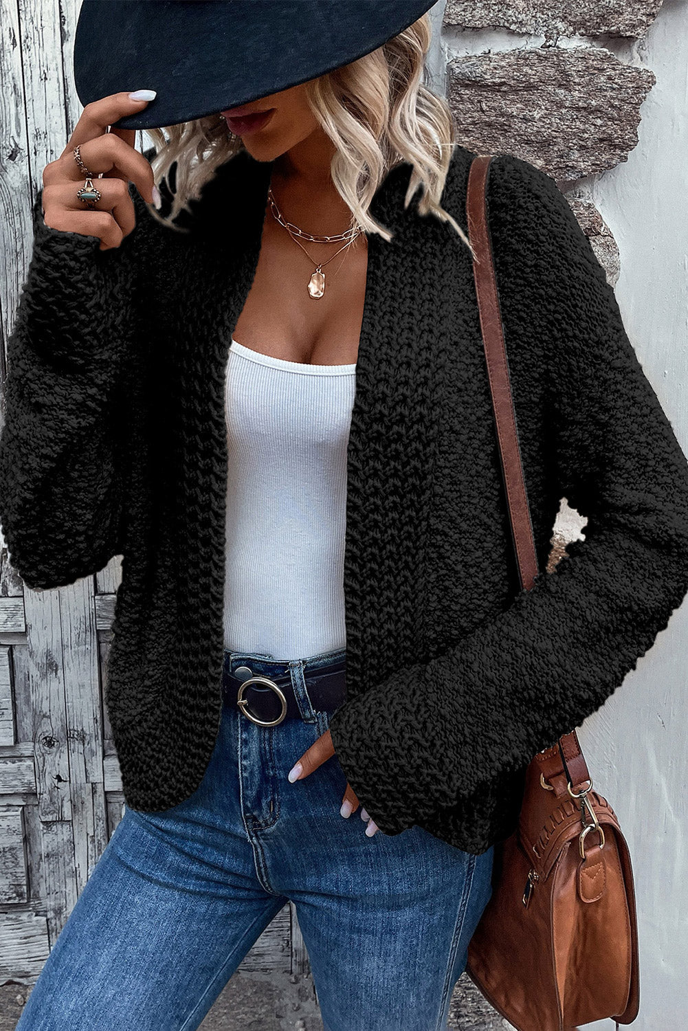 A woman in an Apricot Popcorn Knit Open Front Cardigan and jeans stands against a brick wall, looking to the side with a slight smile.