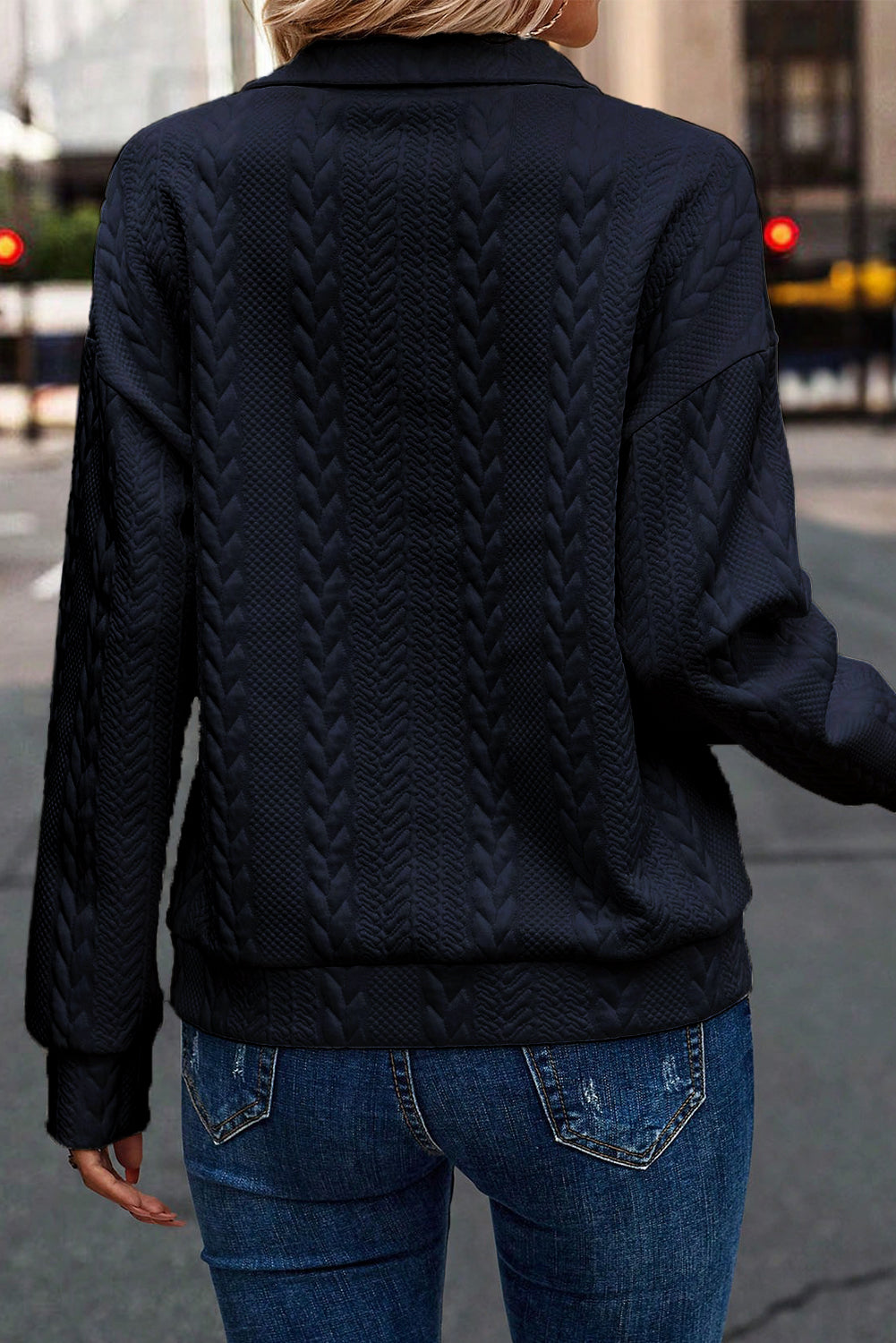 A person wearing a Navy Blue Zip Up Cable Textured Sweatshirt and blue jeans, seen from the back, standing on an urban street.