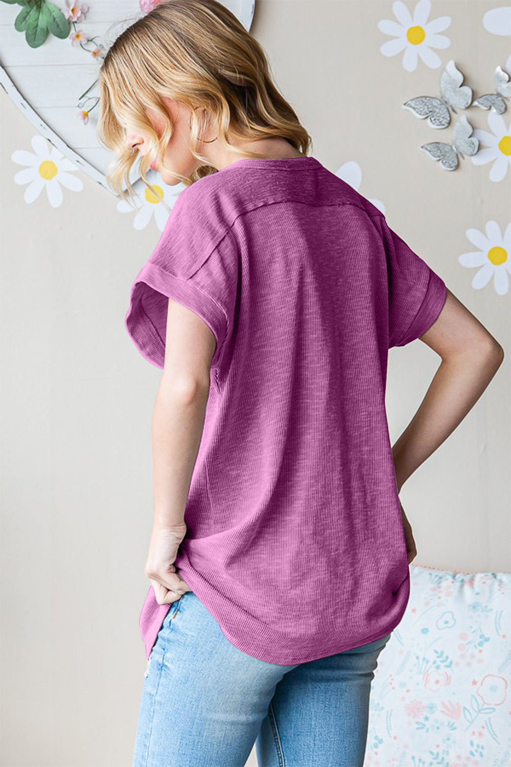 A woman stands in front of a light-colored wall adorned with heart-shaped decor, wearing a pink Heimish Full Size Short Sleeve Round Neck T-Shirt and distressed blue jeans, posing with one hand in her pocket. She has wavy blonde hair. This versatile essential adds a touch of charm to her look.