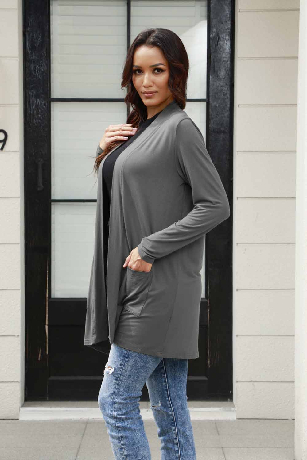 A person with long hair wears the Basic Bae Full Size Open Front Long Sleeve Cardigan with Pockets in gray, paired with a black shirt and blue jeans in a classic basic style. They stand in front of a building's glass door. The ensemble, made from 100% polyester, is perfect for those who prefer machine wash cold for easy care.