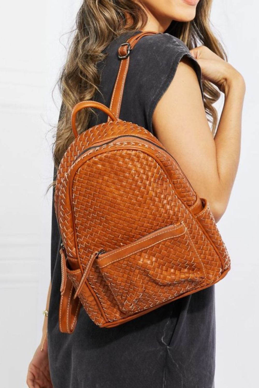 A person wearing the SHOMICO Certainly Chic Faux Leather Woven Backpack over one shoulder, with long hair and a dark shirt, standing sideways.