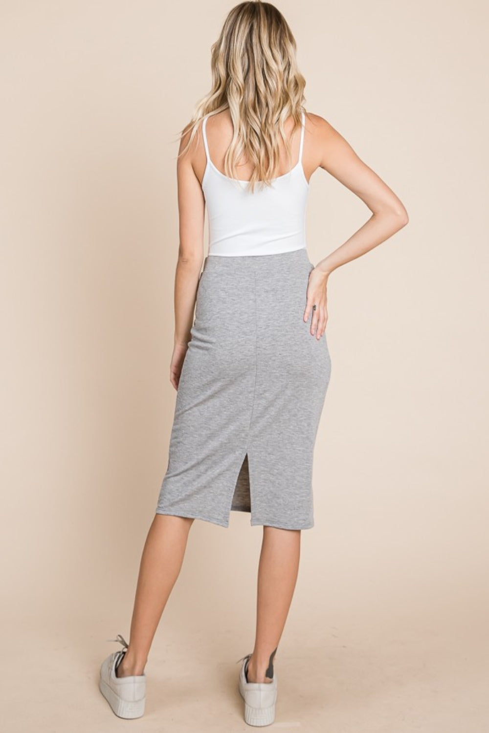 A woman stands against a beige background, wearing a white tank top, the BOMBOM Elastic Waist Pencil Skirt in chic gray made of stretchy fabric and knee-length, paired with white sneakers. She has long blonde hair and a neutral expression.