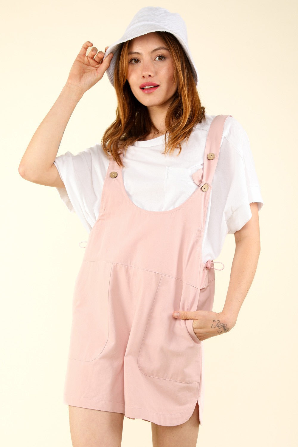 Person wearing a white hat, a white T-shirt, and VERY J Adjustable Waist Suspender Overalls with Pockets in light pink, poses with hands in the side pockets against a plain background.