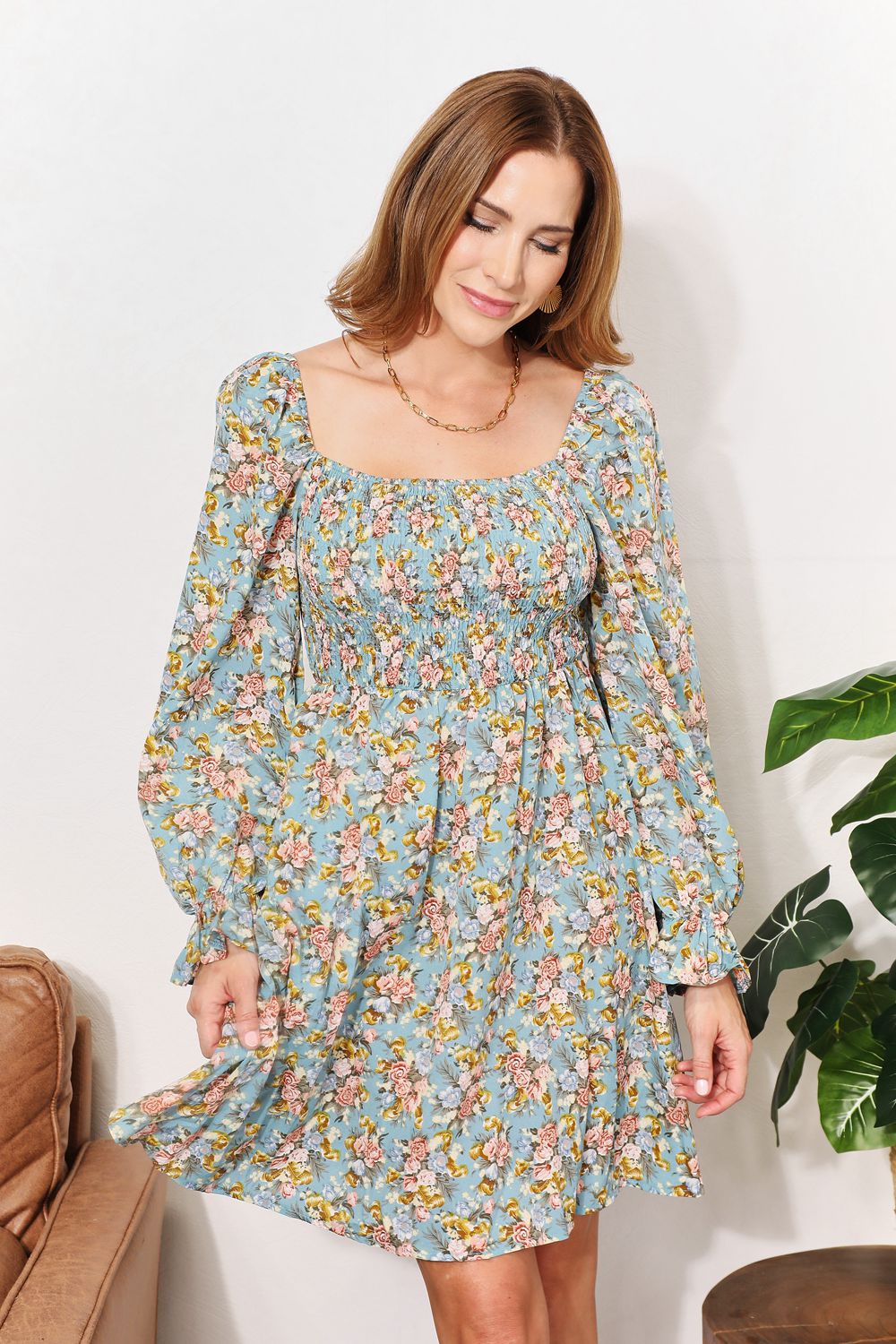 A woman in the Honey Floral Smocked Flounce Sleeve Square Neck Dress stands indoors, holding the hem of her dress. She is smiling slightly with her eyes looking downward. Nearby, a plant and a brown couch complete the cozy scene.