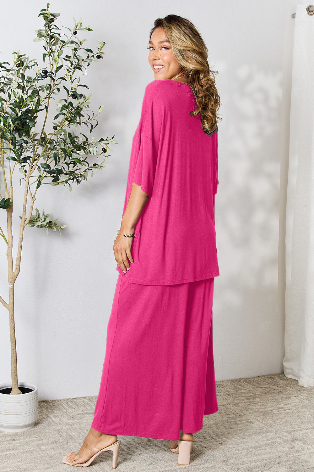 A woman stands near a potted plant and wooden ladder, wearing the Double Take Full Size Round Neck Slit Top and Pants Set in a bright pink two-piece outfit with matching wide-leg pants that are available in plus size and are easily machine washable.