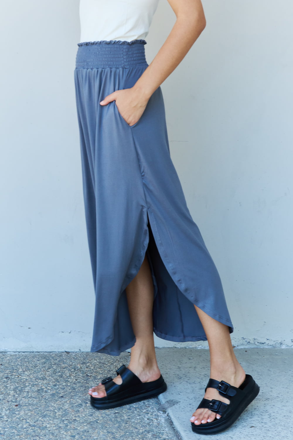 A person wearing a white sleeveless top and the Doublju Comfort Princess Full Size High Waist Scoop Hem Maxi Skirt in Charcoal stands against a plain wall. They are also wearing black sandals.