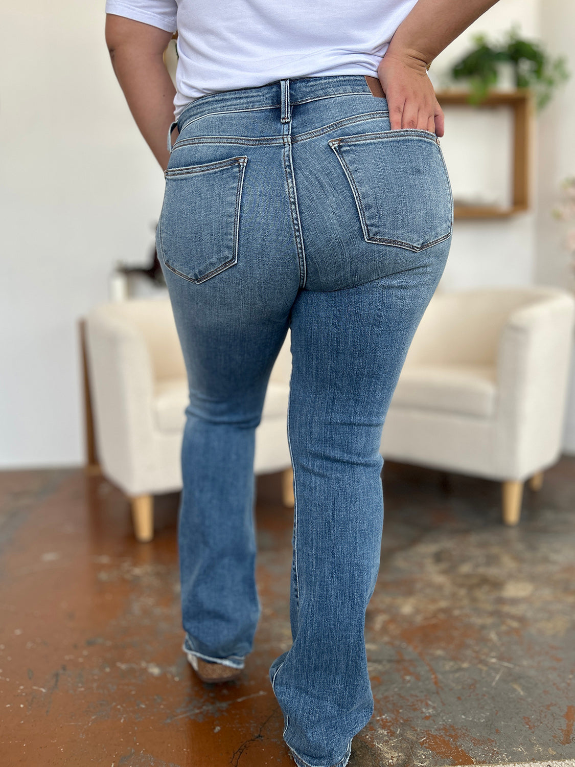 Person wearing Judy Blue Full Size Mid-Rise Waist Straight Jeans and a black top, standing in a room with white chairs, potted plants, and wooden shelves. Only the lower half of the body and part of the arms are visible.