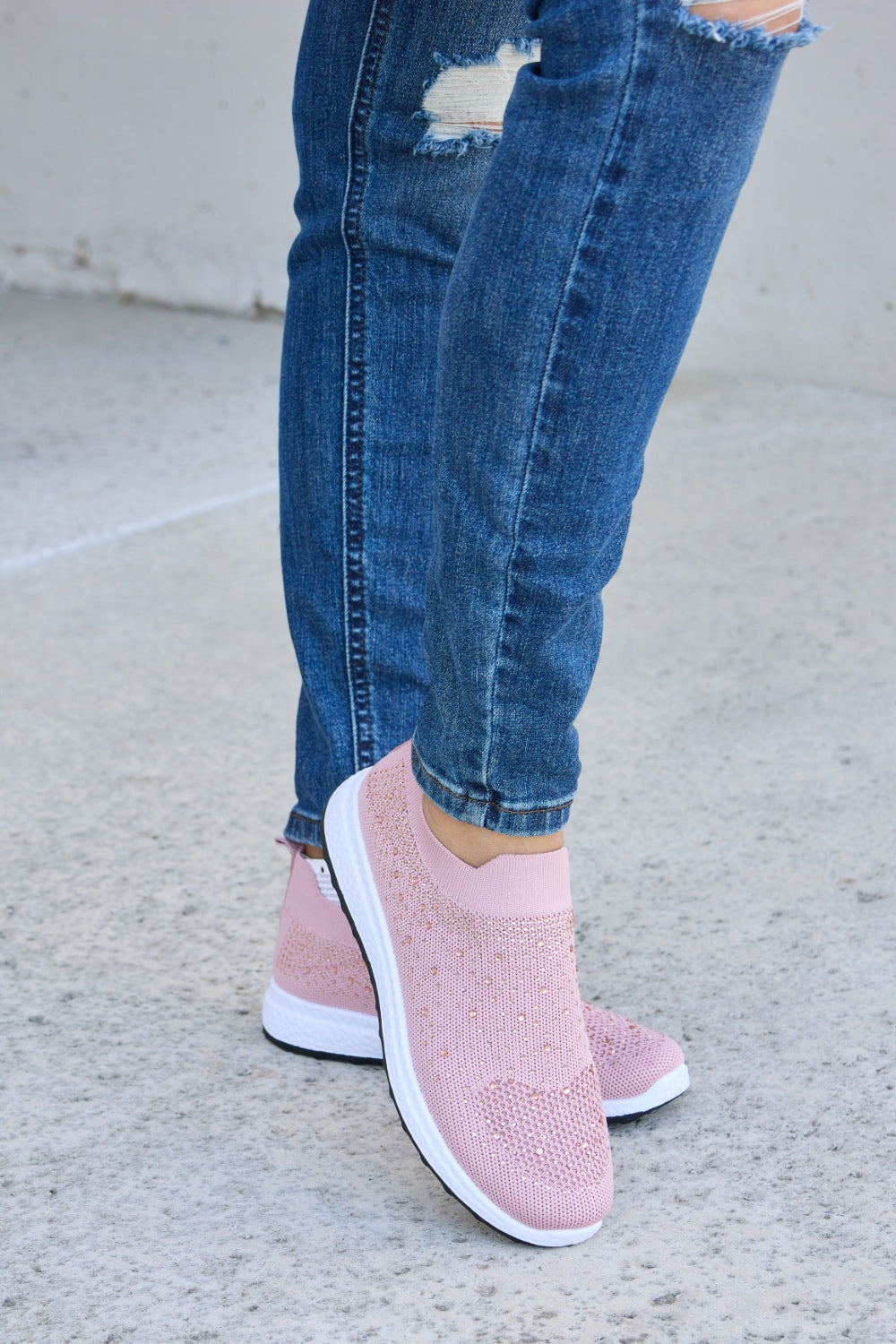 Person wearing Forever Link Rhinestone Mesh Breathable Sneakers in light pink and ripped blue jeans, standing on a concrete surface.