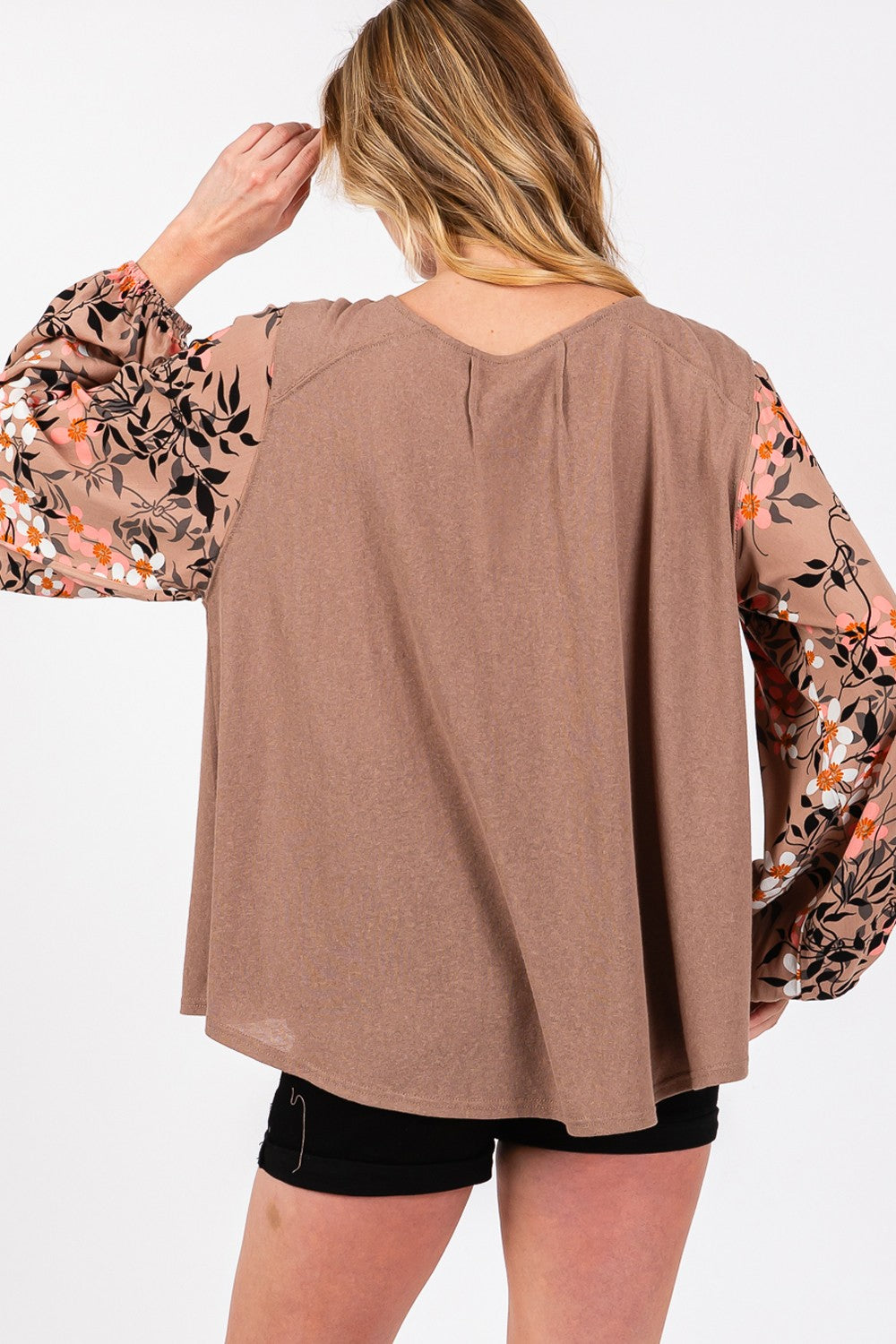 A person is wearing the SAGE + FIG Floral Long Sleeve Front Pleated Detail Blouse, paired with black shorts and a black hat, standing against a plain background.