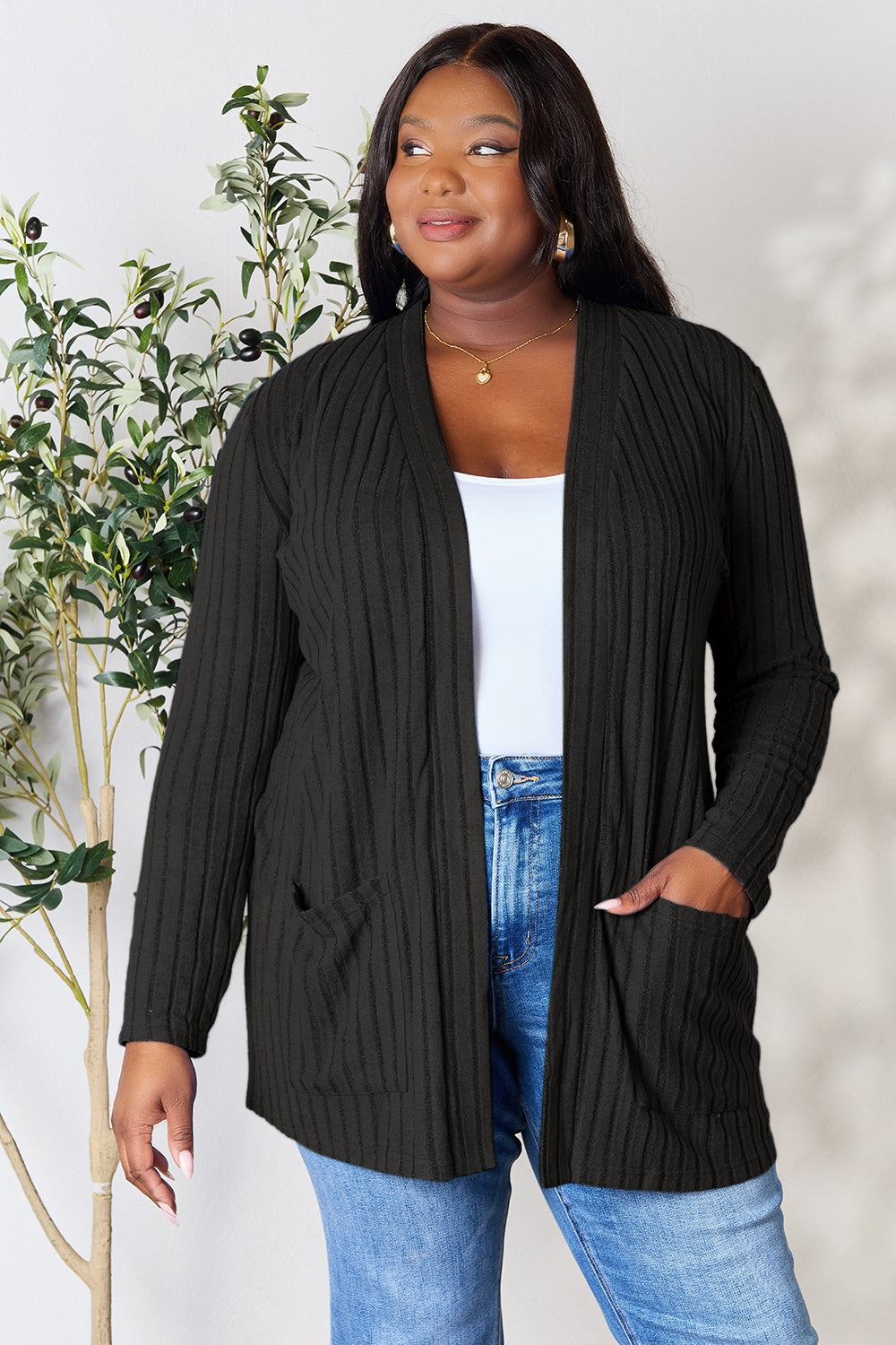 A woman in a Basic Bae Full Size Ribbed Open Front Cardigan with Pockets and jeans, made from stretchy material, stands in front of a plant, smiling slightly and looking to the side.