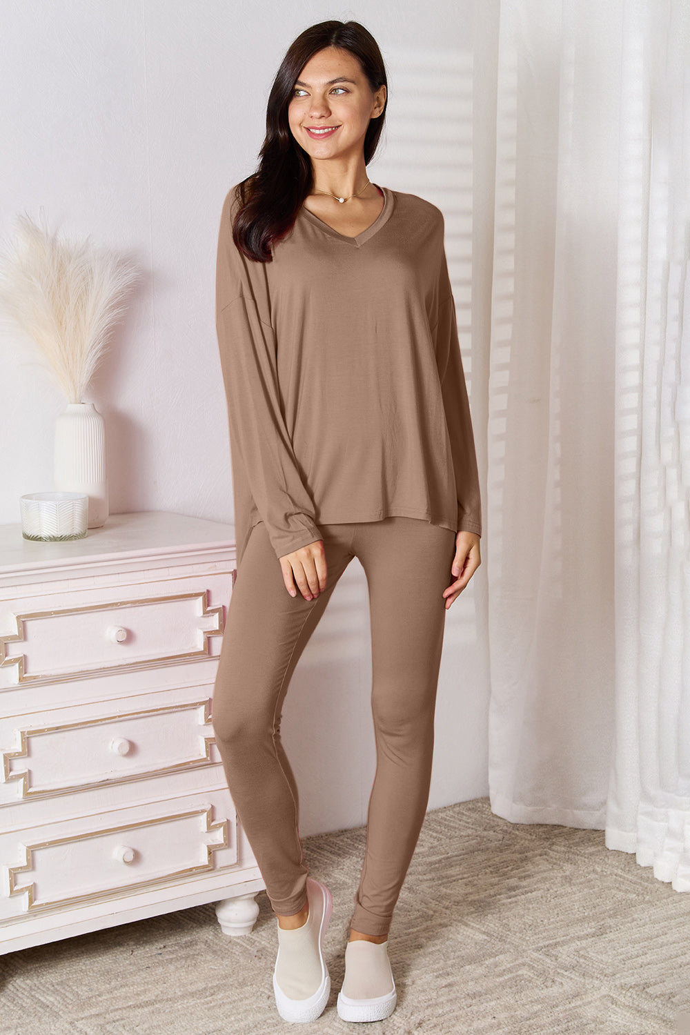 A woman stands indoors, posing in front of a white dresser next to a tall vase with decorative white feathers. She is wearing the Basic Bae Full Size V-Neck Soft Rayon Long Sleeve Top and Pants Lounge Set in bright pink, paired with highly stretchy material and completed with white slip-on shoes.