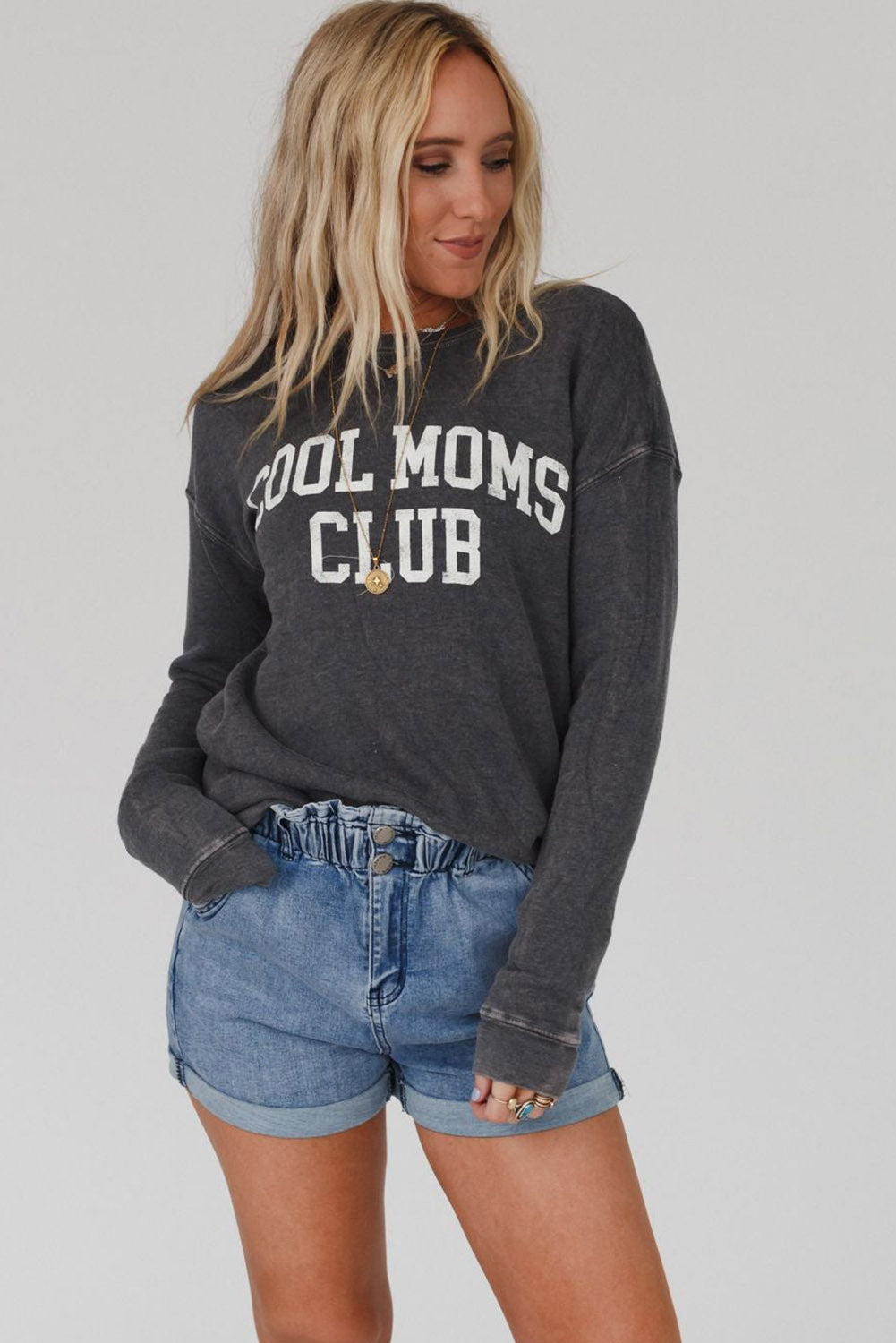 A person with long blond hair wearing a gray COOL MOMS CLUB drop shoulder sweatshirt and relaxed-fit blue jeans is seen from the back, standing near a pumpkin and a window.