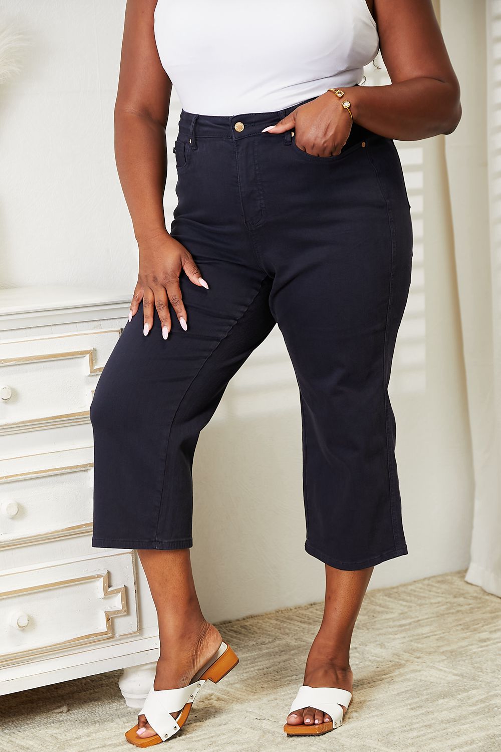 A person wearing a white sleeveless top and Judy Blue Full Size High Waist Tummy Control garment-dyed wide cropped jeans in black stands indoors next to a white dresser.