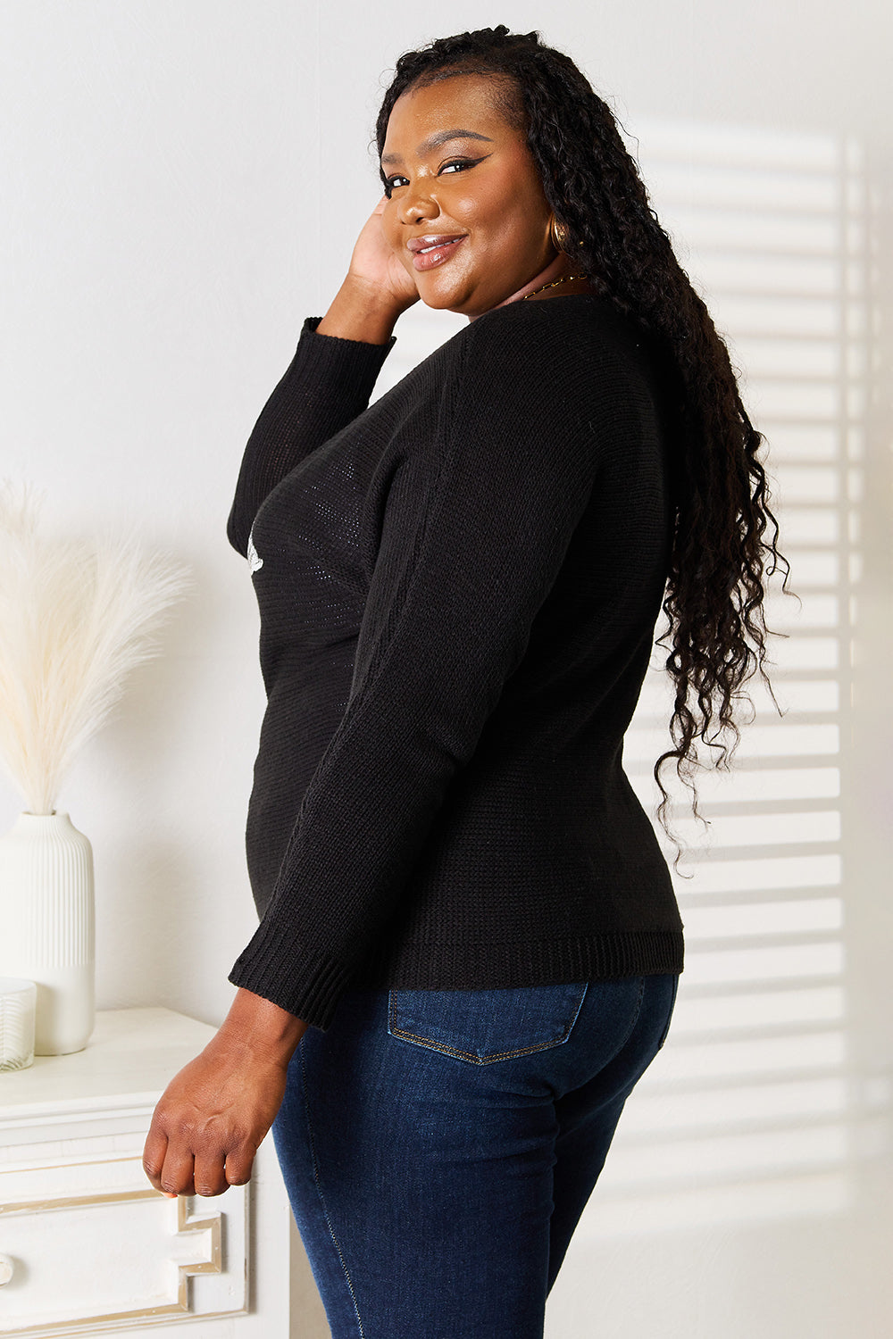 A person wearing a Sequin Graphic Dolman Sleeve Knit Top paired with blue jeans stands near a white dresser adorned with decor items.