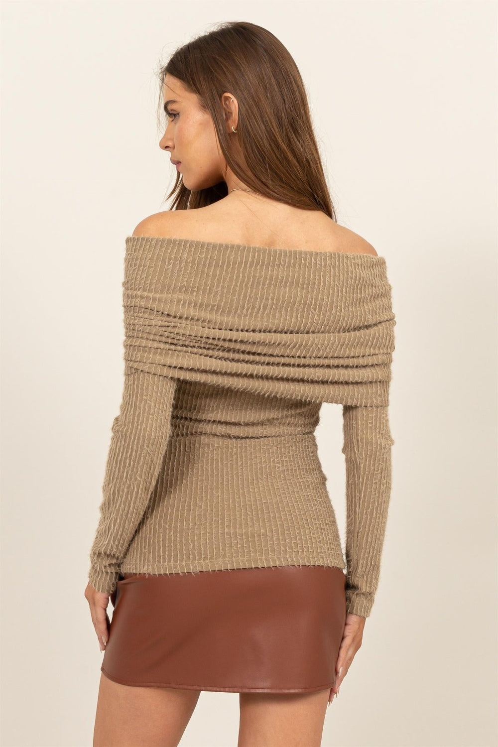 A woman elegantly stands against a light background, wearing the chic HYFVE Fuzzy Off Shoulder Textured Knit Top paired with a brown leather skirt.