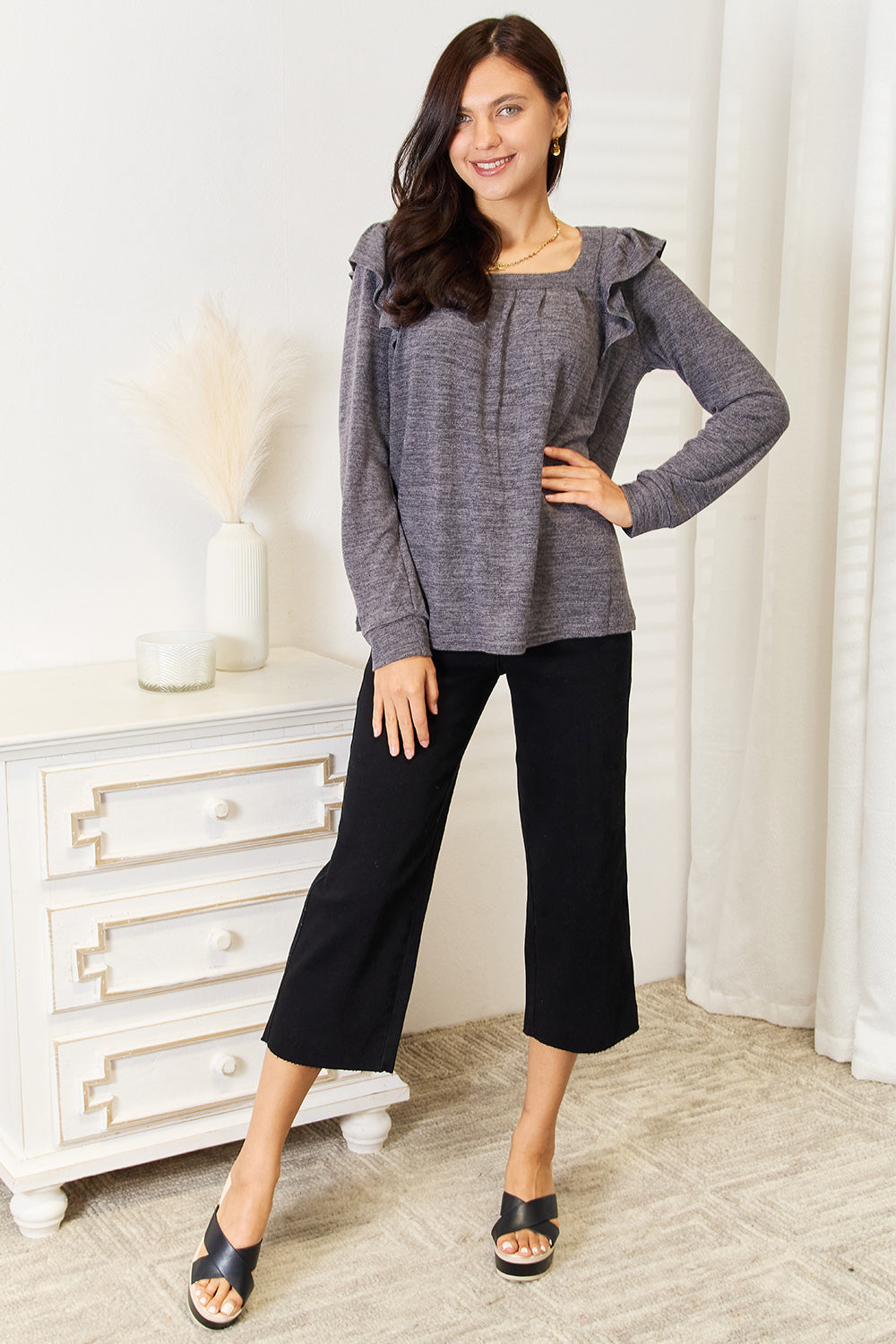 A woman wearing a Double Take Square Neck Ruffle Shoulder Long Sleeve T-Shirt in pink and blue jeans stands with one hand in her pocket, smiling. She has long curly hair and wears a necklace and bracelet. She stands in a room with white decor.