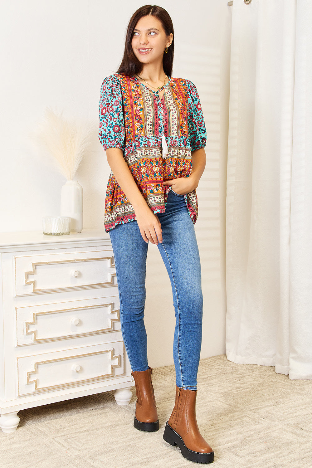 A woman wearing the Devine Bohemian Tassel Tie Puff Sleeve Babydoll Blouse, adorned with a mix of vibrant floral designs, smiles in her jeans while standing indoors against a light background.