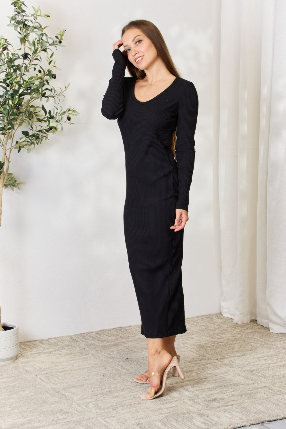 A person stands indoors wearing the chic Culture Code Full Size Ribbed Long Sleeve Midi Slit Dress paired with sandals, next to a potted plant.