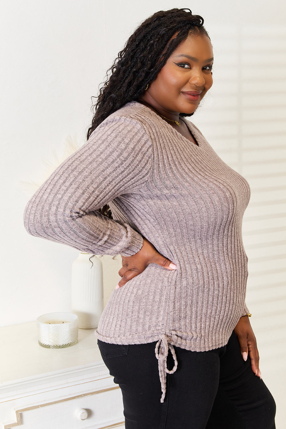 A woman stands indoors against a neutral background, wearing the "Double Take Drawstring Ribbed Long Sleeve T-Shirt" in light purple paired with dark pants. She is smiling and posing with one hand on her hip.