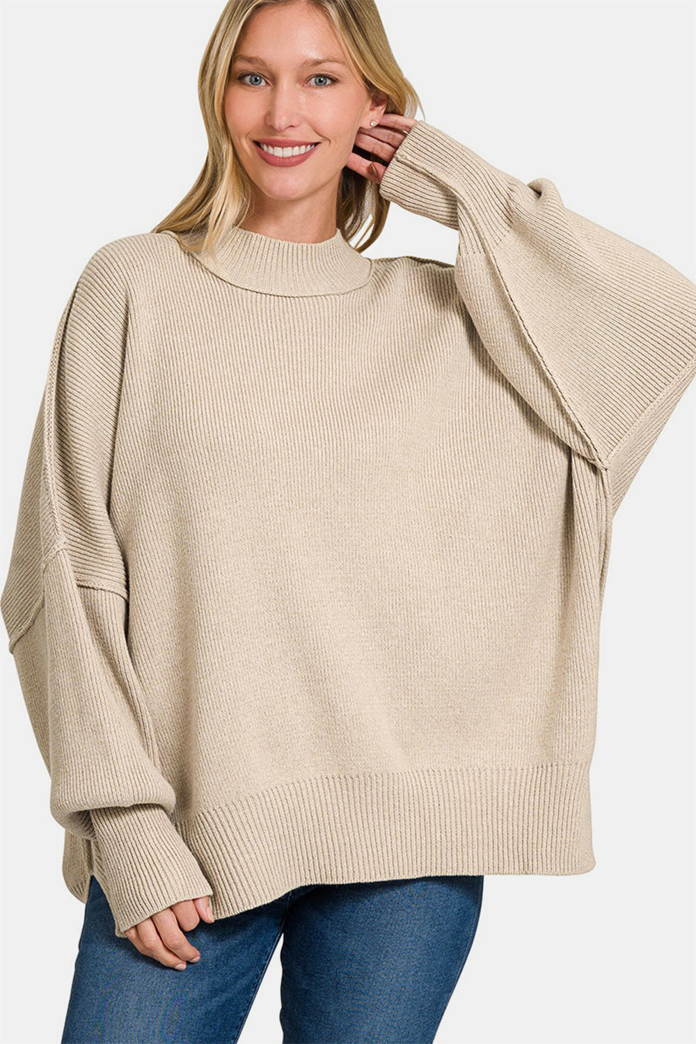 A person wearing the Zenana Side Slit Oversize Sweater, a cozy knit in beige, paired with jeans, smiling with one hand touching their hair.