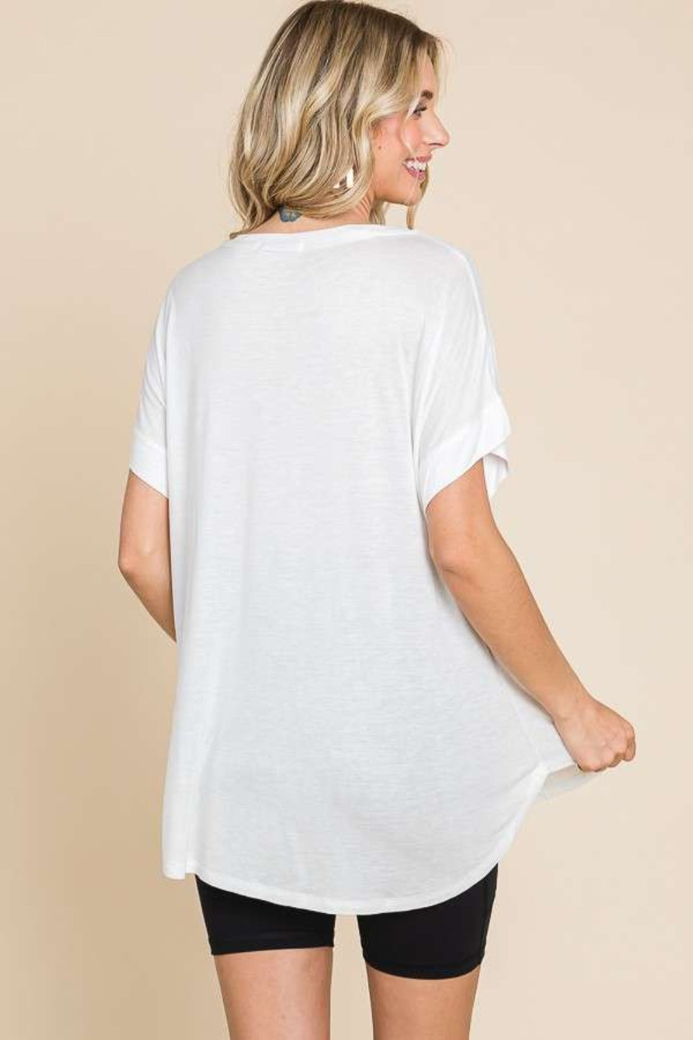 Woman in a white Culture Code Round Neck Short Sleeve T-Shirt and black shorts, posing against a beige background with one hand on her hip. This versatile staple makes everyday wear effortlessly chic.