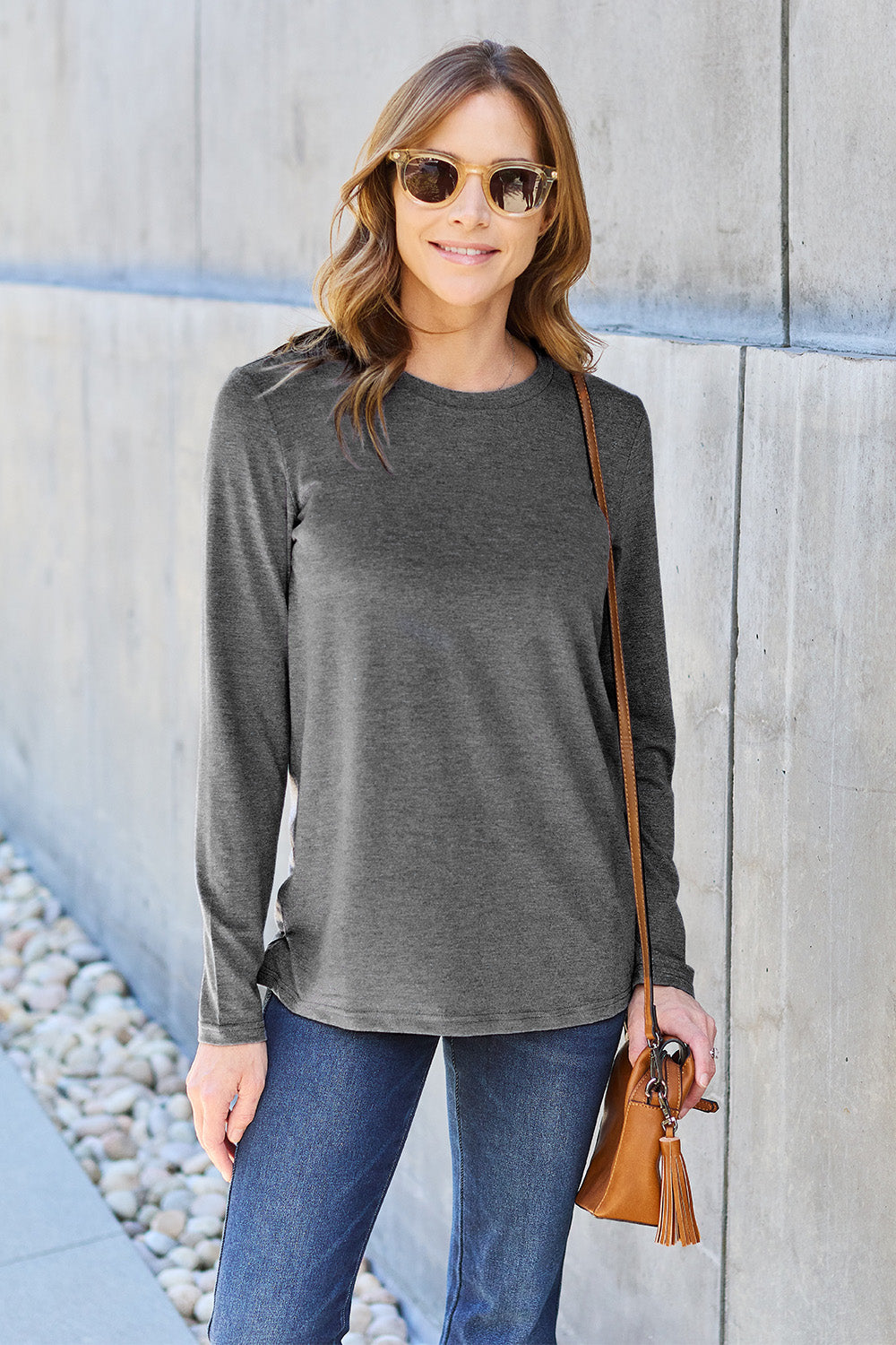 A woman with shoulder-length hair wearing the Basic Bae Full Size Round Neck Long Sleeve Top and blue jeans stands in front of a concrete wall, holding a beige handbag over her shoulder. Her outfit exemplifies basic style, with the top made from 100% polyester and recommended for machine wash cold.