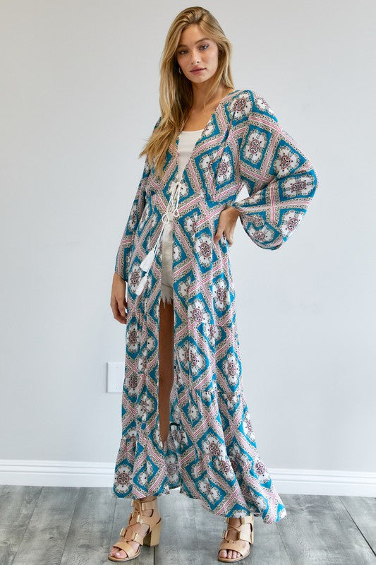 A person wearing a Printed Long Sleeve Loose Kimono with a bohemian blue and pink print over a white outfit stands in a room with a gray floor and white wall.