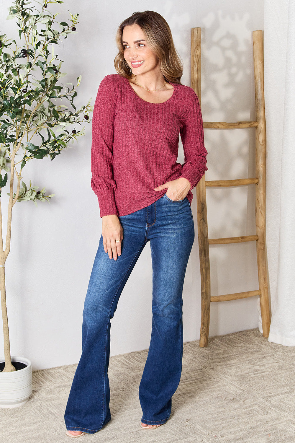 A person in a stylish maroon Ribbed Round Neck Lantern Sleeve Blouse and jeans stands by a wooden ladder, with olive branches in the background.