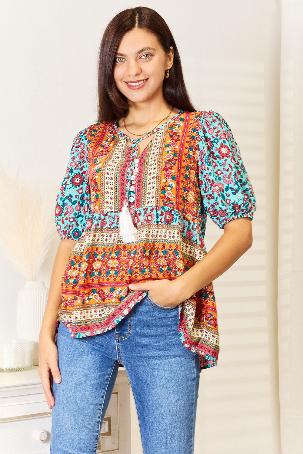 A woman wearing the Devine Bohemian Tassel Tie Puff Sleeve Babydoll Blouse, adorned with a mix of vibrant floral designs, smiles in her jeans while standing indoors against a light background.