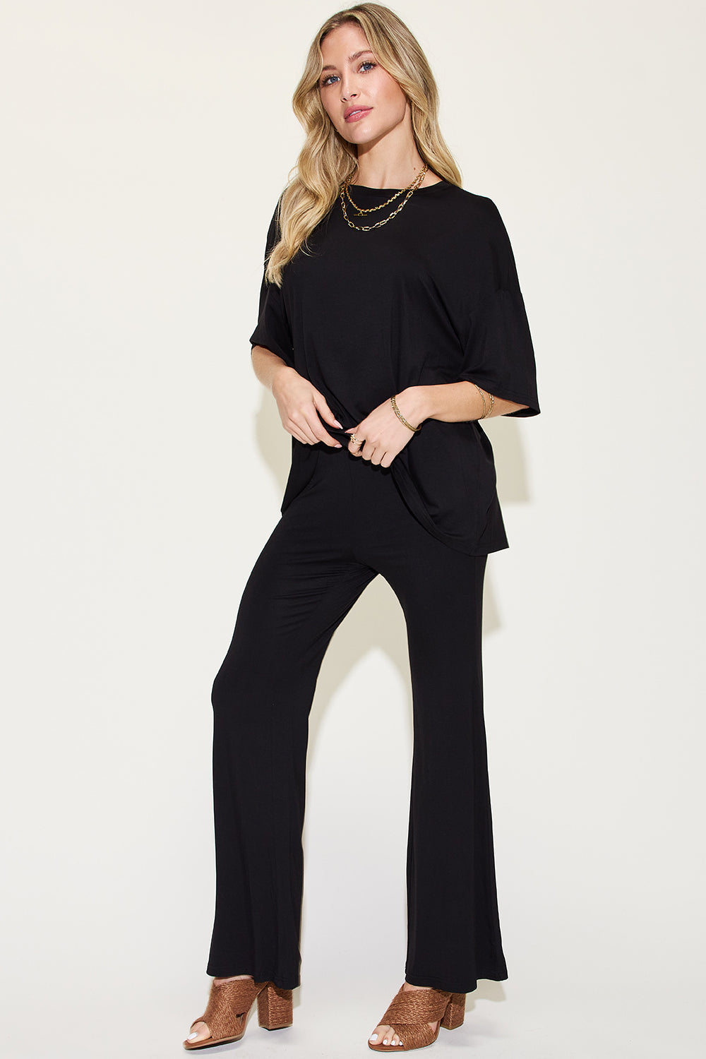 A woman with long, blonde hair wears the Basic Bae Full Size Bamboo Drop Shoulder T-Shirt and Flare Pants Set in black, paired with brown heeled sandals and layered necklaces. She stands against a plain, light background.